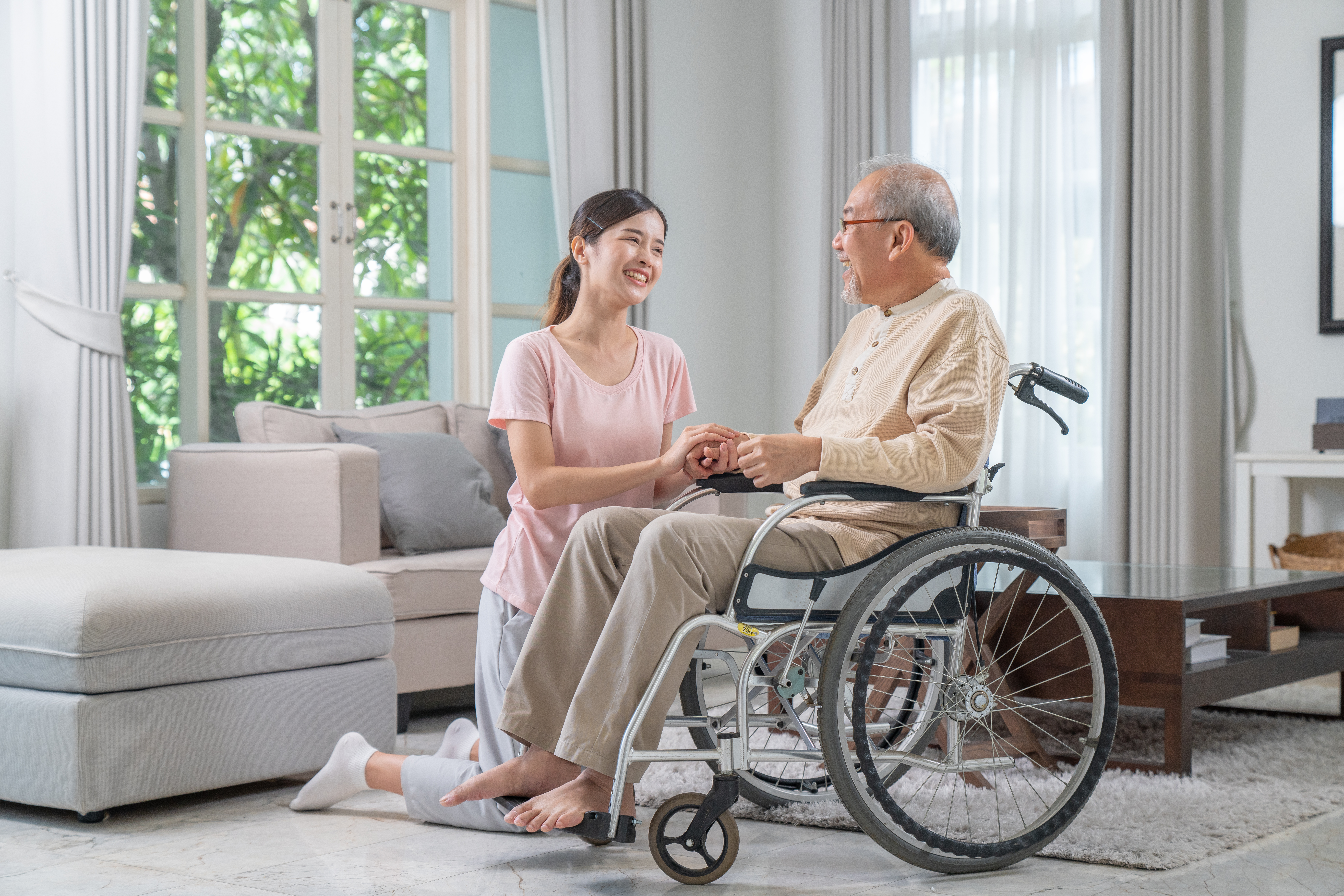 A young woman caring for her grandfather in a wheelchair, smiling warmly while holding his hands in a bright living room.