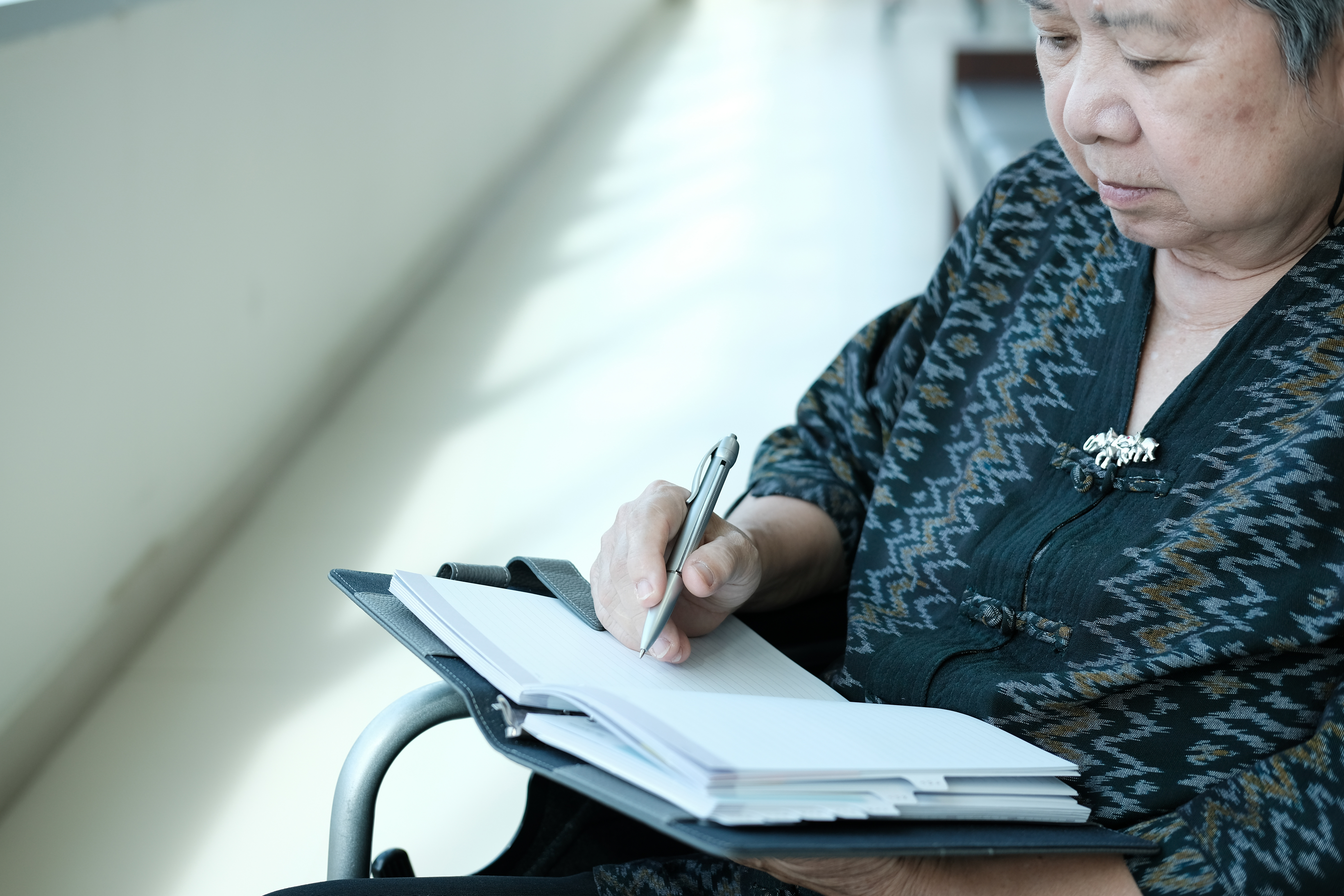 An elderly woman sitting in a wheelchair, thoughtfully writing in a notebook while preparing to draft her will.